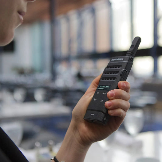 A person holding a black Motorola walkie-talkie in a real indoor environment with a blurred office or meeting room background.