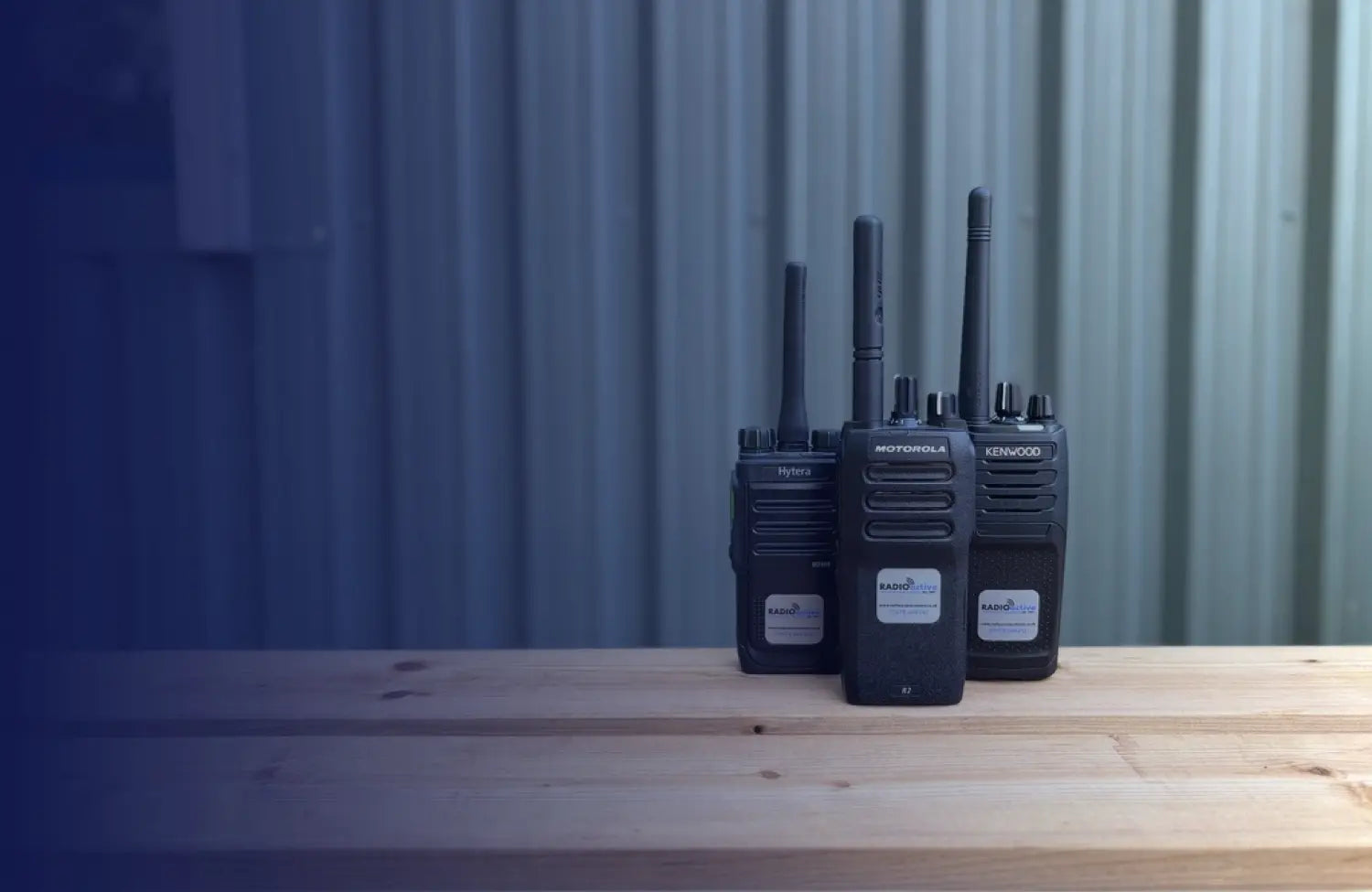 Three black Motorola handheld walkie-talkies standing upright on a light wooden surface against a corrugated metal wall.