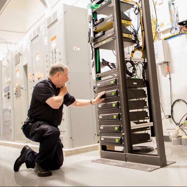 A male technician in a black uniform kneels beside a tall server rack filled with networking equipment inside a clean data center with white walls, cables, and other server cabinets.