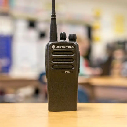 A black Motorola walkie-talkie standing upright on a light wood surface with a blurred indoor background.