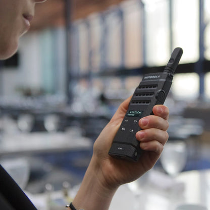 A person holding a black Motorola walkie-talkie in a real indoor environment with a blurred office or meeting room background.