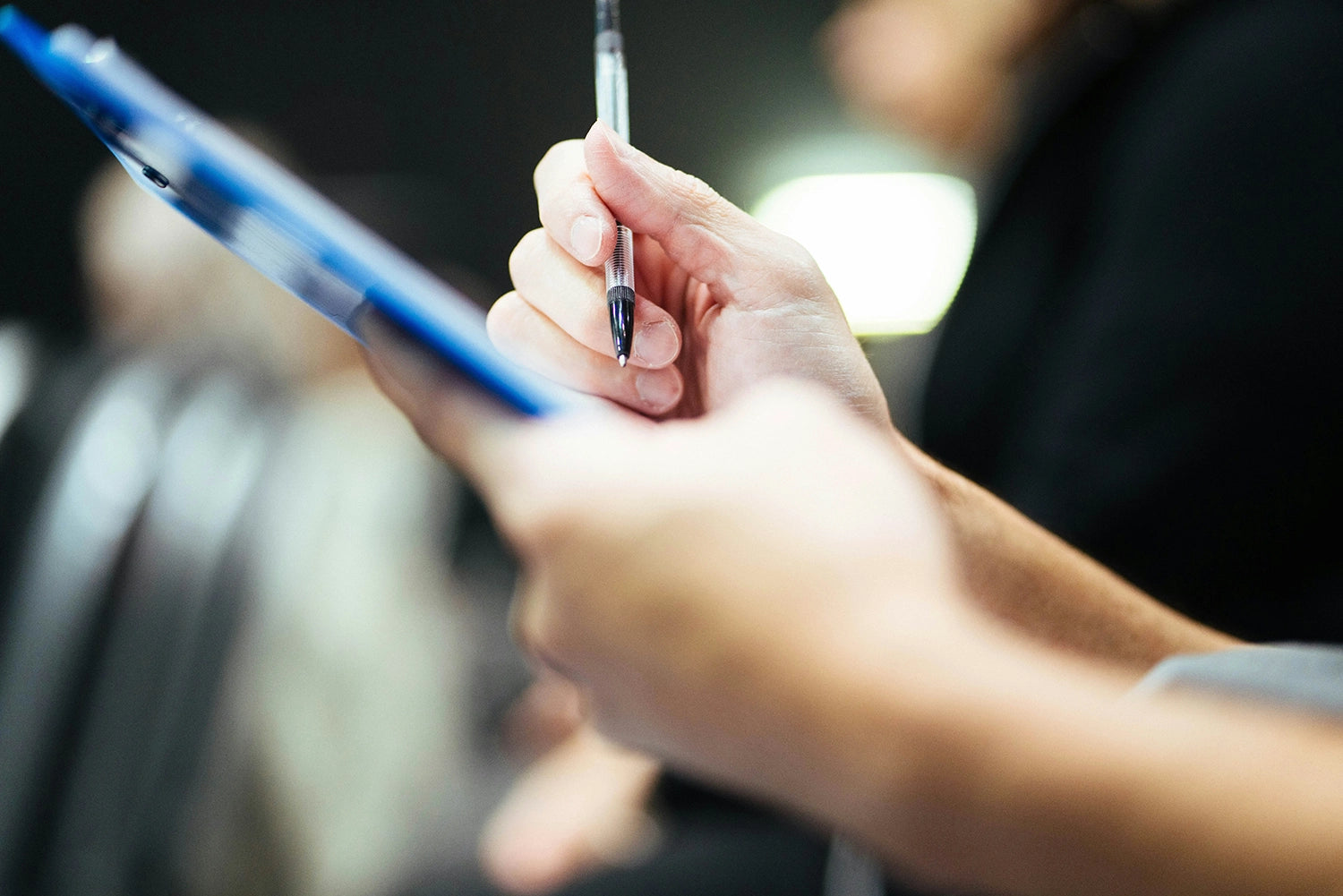 A close-up shot of a person's hand holding a black pen and a blue clipboard, captured in a softly blurred indoor setting.